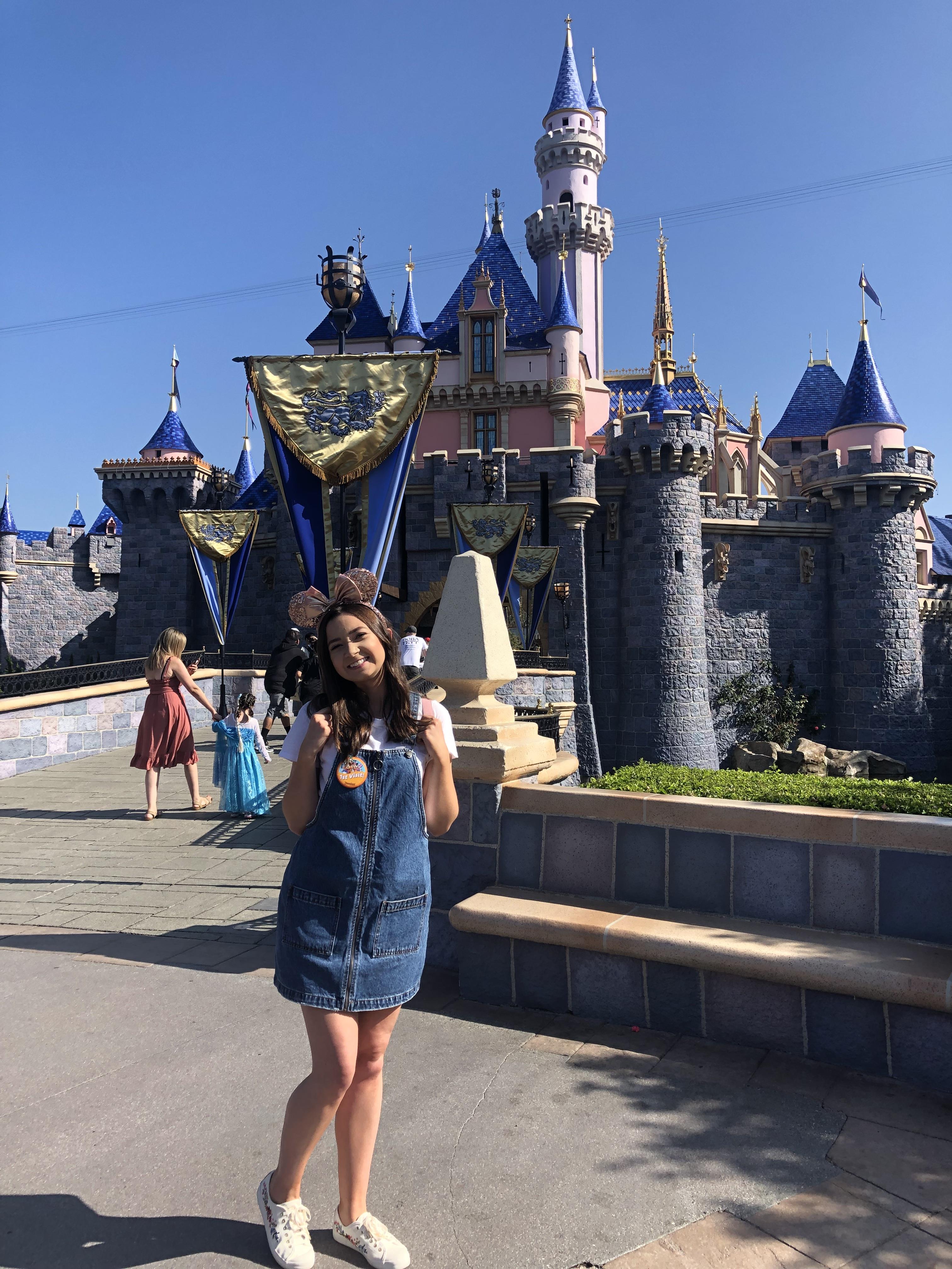 A young woman (the author, Cami) wearing overalls dress and pink Mickey Mouse ears, poses in front of a castle at Disneyland.