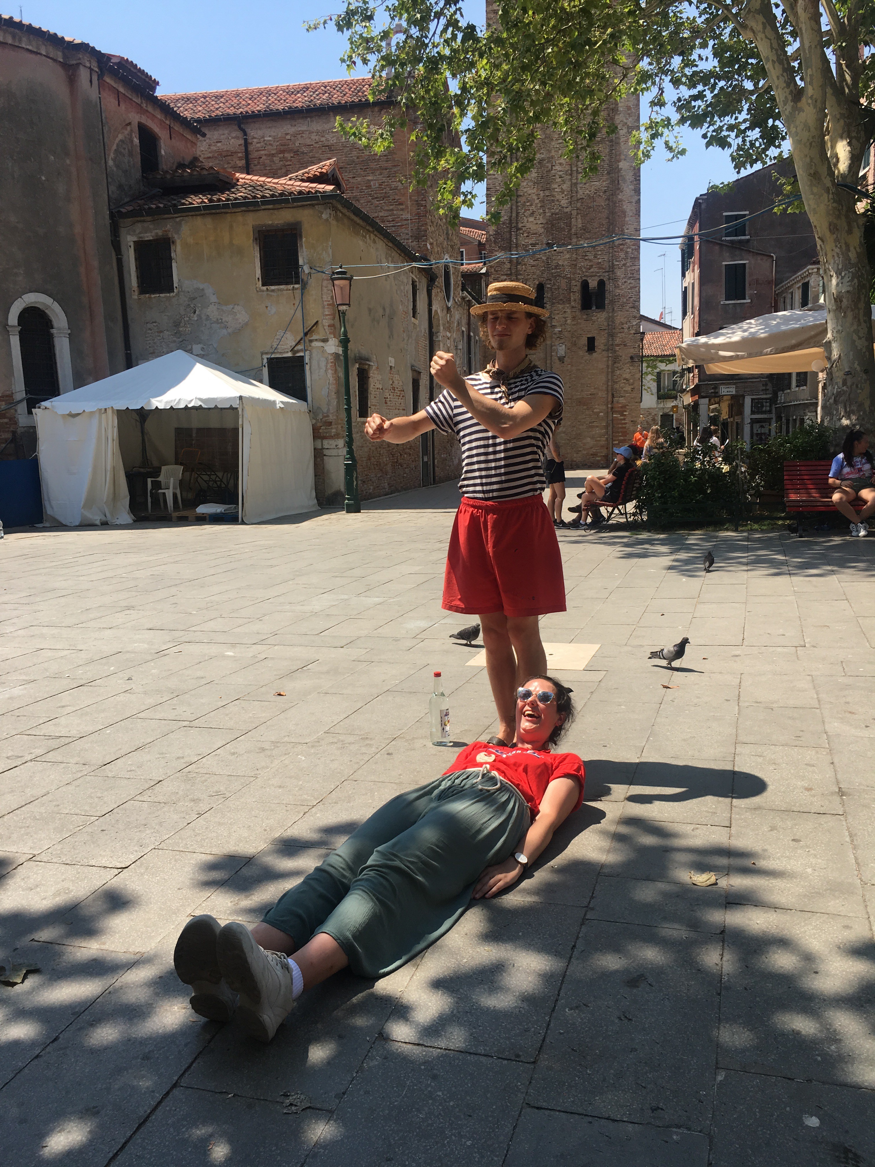 On a street in Venice, a young woman lies on the ground pretending to be a gondola while a gondolier stands over her, pretending to paddle.