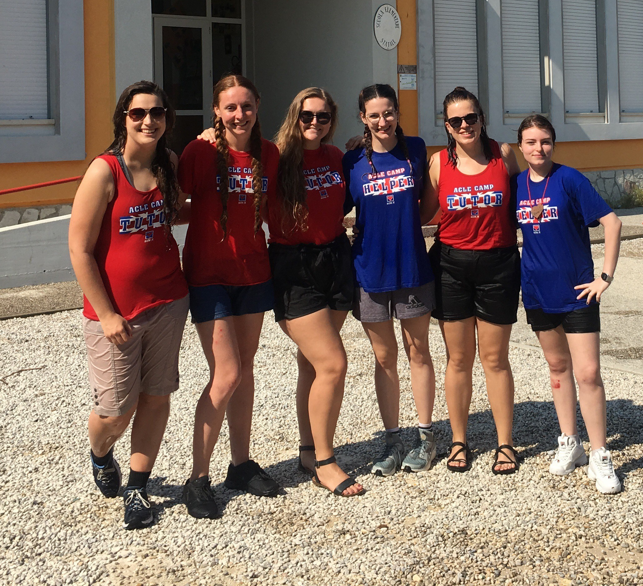 Six young women pose outside in a row, smiling and wearing red or blue shirts that read "ACLE CAMP TUTOR."