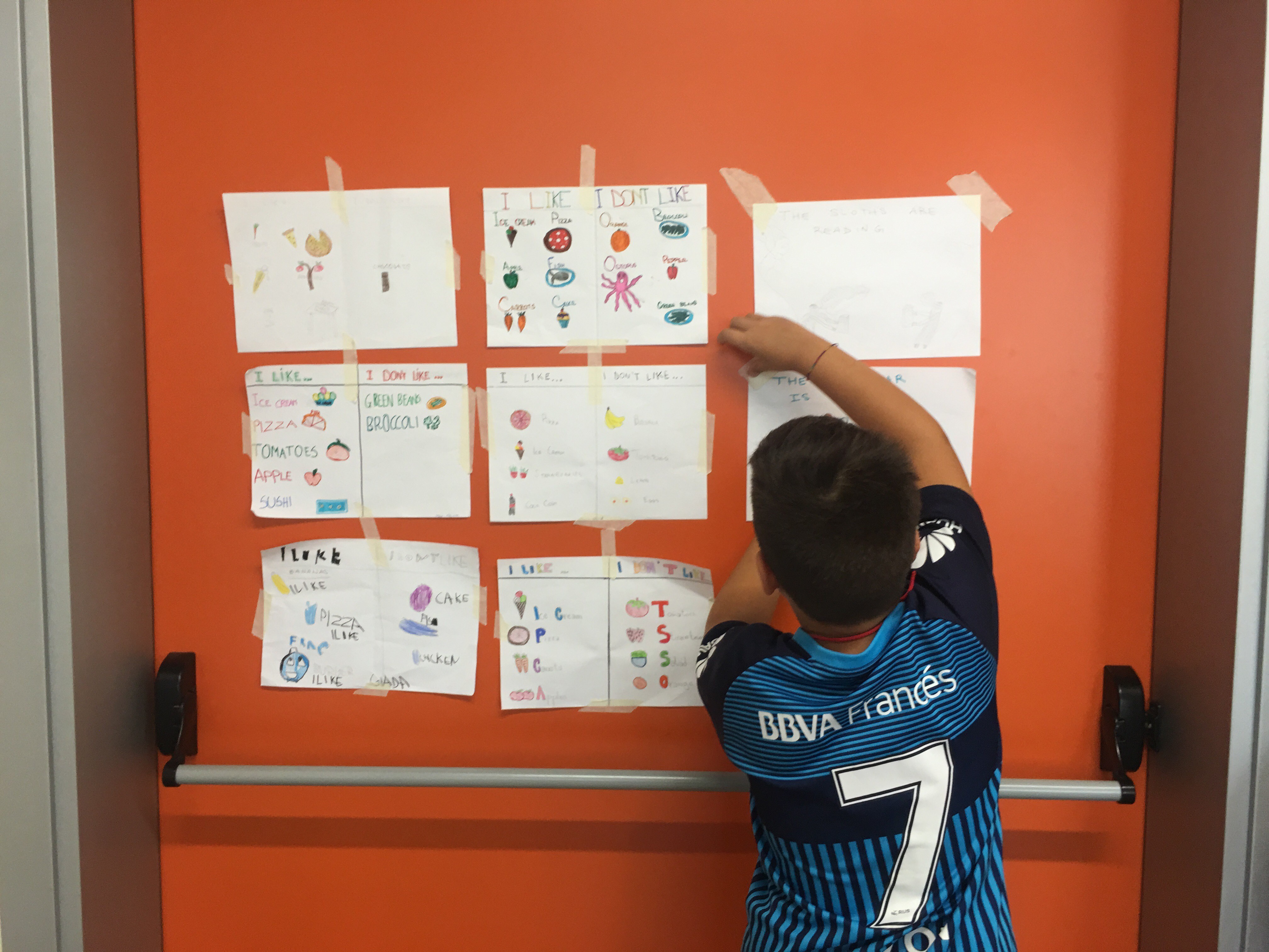 A young boy with his back to the camera posts a drawing on an orange wall next to several other drawings.