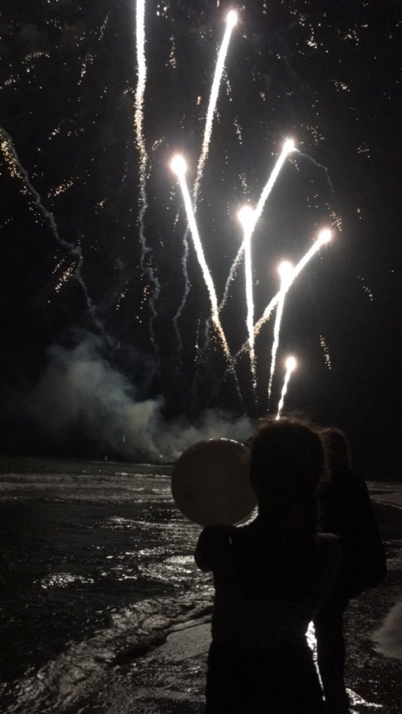 Image shows fireworks and white balloons at a beach at night time. The photo is dark and the fireworks are bright.