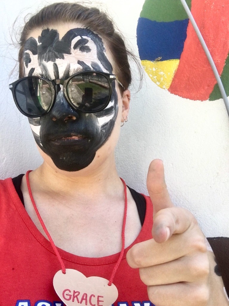 A young woman poses for the camera, wearing sunglasses and face paint that resembles a zebra for a camp activity. She points a finger gun at the camera to look cool.